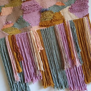 May include: A woven wall hanging with a geometric pattern in shades of pink, green, and gold. The hanging has long fringe tassels in the same colors.
