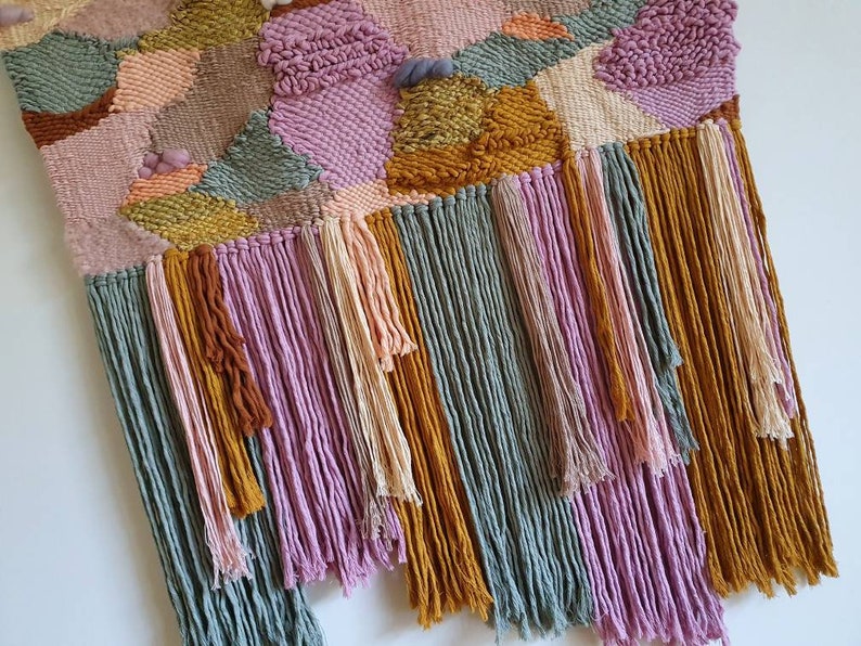 May include: A woven wall hanging with a geometric pattern in shades of pink, green, and gold. The hanging has long fringe tassels in the same colors.