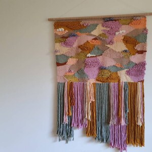 May include: A colorful woven wall hanging with a geometric pattern in shades of pink, green, yellow, and brown. The hanging has a fringe border and is made with a variety of textures.