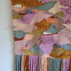 May include: A colorful woven wall hanging with a variety of textures and patterns. The hanging features a mix of pink, green, yellow, and brown yarn, and has a fringe border.