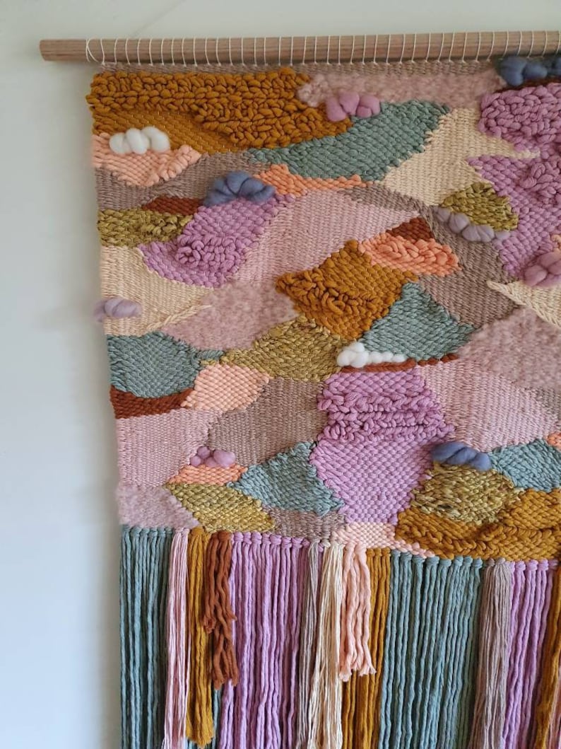 May include: A colorful woven wall hanging with a variety of textures and patterns. The hanging features a mix of pink, green, yellow, and brown yarn, and has a fringe border.