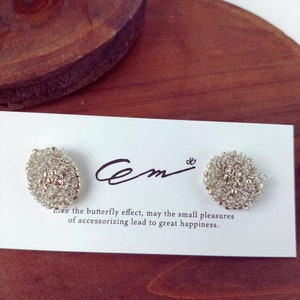 May include: A pair of silver beaded earrings on a white card with the text "Cem" and "Like the butterfly effect, may the small pleasures of accessorizing lead to great happiness."