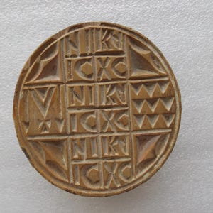 May include: A round, wooden carving with a geometric design. The carving features a grid pattern with the Greek letters NIKA and ICXC. The outer edge has a decorative border. The wood has a warm, light brown color.