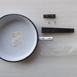 Może przedstawiać: Biała patelnia emaliowana z niebieską obwódką, wraz z odłączonym czarnym uchwytem, osprzętem montażowym i plastikową torbą. Patelnia ma etykietę w środku. Uchwyt ma około 18 cm długości.
