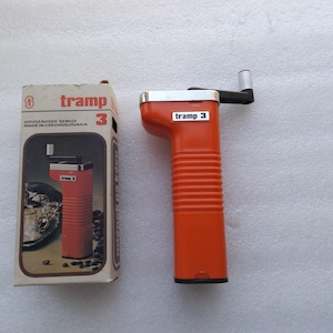 May include: An orange hand-crank coffee grinder with the text "tramp 3" on the grinder and the box. The box is beige with the text "tramp 3" and an image of the grinder. The grinder has a black handle and a silver top.