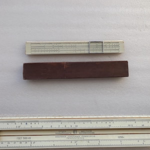 May include: A vintage slide rule with a cream-coloured body and black markings, alongside a brown wooden case. The slide rule features a clear cursor and various scales for calculations. The case is rectangular and appears to be made of dark brown wood.