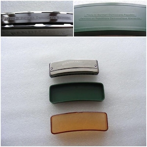 Pode incluir: Uma gaita de boca prateada com as palavras "BANDMASTER SUPER" impressas na frente. A gaita de boca está em uma caixa de plástico verde com as palavras "Made in German Democratic Republic" impressas na parte superior. A gaita de boca também está em uma caixa de plástico marrom.