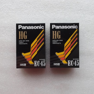 Könnte beinhalten: Zwei Panasonic HG VHS-Videokassetten. Die schwarzen Boxen tragen das Panasonic-Logo und den Text "HG SUPER HIGH GRADE COMPACT VIDEOCASSETTE". Die Boxen haben ein Design aus orangefarbenen, gelben und roten Linien. Die Modellnummer ist EC-45.