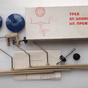 May include: A white box with the text "УРЕД ЗА НАВИВАНЕ НА ПРЕЖДА" in red letters. The box contains a metal tool for winding yarn. The tool has two blue plastic spools, a metal clamp, and a metal rod. There are also several metal rods and a small instruction sheet.