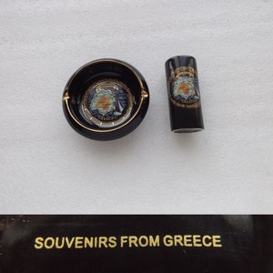 Pode incluir: Cinzeiro de cerâmica preto e recipiente cilíndrico correspondente com guarnição dourada e um mapa da Grécia com a palavra "Grécia" impressa em ambos. O texto "Souvenirs from Greece" está impresso em dourado sobre um fundo preto.