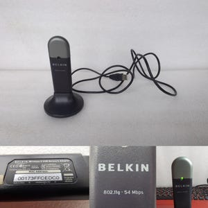 Może przedstawiać: Czarny adapter sieciowy USB Belkin Wireless G z kablem USB. Adapter ma zaokrągloną górę i okrągłą podstawę. Tekst "BELKIN" jest widoczny na urządzeniu. Jest przeznaczony do łączności sieci bezprzewodowej.