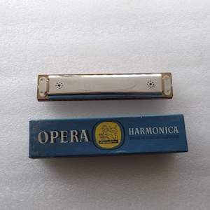 Pode incluir: Uma gaita vintage com corpo prateado e caixa azul. A caixa diz "OPERA HARMONICA MADE IN CZECHOSLOVAKIA" com um emblema amarelo. A gaita é um instrumento musical.