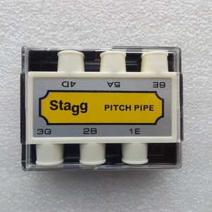 May include: A white plastic pitch pipe set with four tuning pipes in a clear plastic case. The case has a silver label with the text "Stagg PITCH PIPE" in black letters and the notes "3G", "2B", "1E", "G4", "A4", and "B5" printed in black.