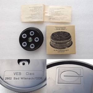 May include: A selection of vintage SKRIMAT items. Includes a black ink pad holder with white rings, a cardboard box, and a metal component with the text "VEB Cleo 2902 Bad Wilsnack/DDR".