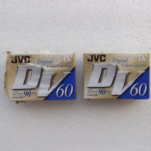 May include: Two JVC Digital DV 60 video cassettes. The packaging is tan with blue accents and silver lettering. The text "JVC", "Digital Video Cassette", "DV", and "60" are visible. The cassettes are labeled LP mode 90 min.