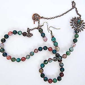 May include: A jewelry set featuring a necklace, bracelet, and earrings. The set includes round beads in shades of green, red, and gray. The necklace and bracelet are connected with gold-colored clasps. The earrings have a hook design.