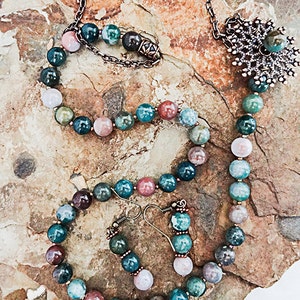 May include: A necklace, bracelet, and earring set featuring multicolored gemstone beads. The necklace has a decorative pendant and a chain. The jewelry is displayed on a textured, light brown surface.