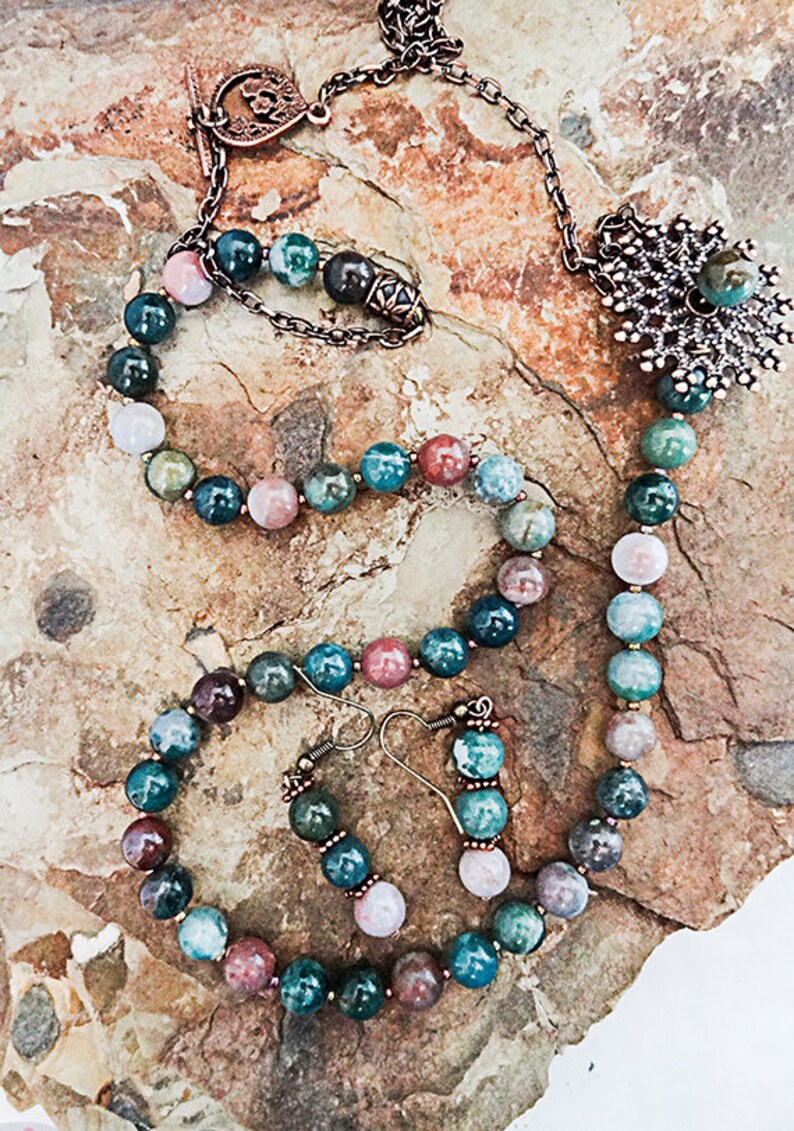 May include: A necklace, bracelet, and earring set featuring multicolored gemstone beads. The necklace has a decorative pendant and a chain. The jewelry is displayed on a textured, light brown surface.