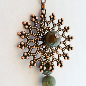 May include: A copper-colored pendant necklace with a snowflake design. The pendant has a central green stone and is connected to a chain. Below the pendant are several green and pink gemstone beads.