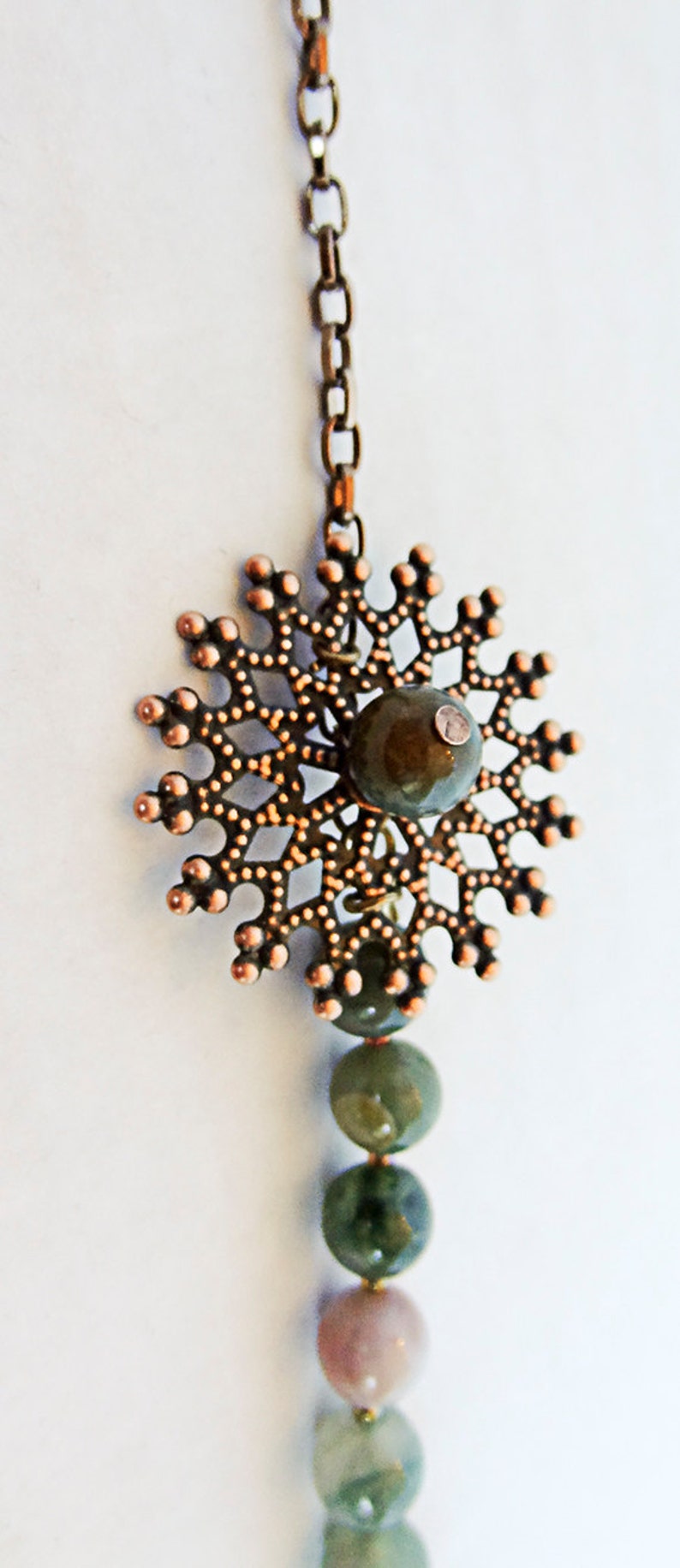May include: A copper-colored pendant necklace with a snowflake design. The pendant has a central green stone and is connected to a chain. Below the pendant are several green and pink gemstone beads.