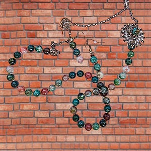 May include: A jewelry set featuring a necklace, bracelet, and earrings with colorful beaded accents. The necklace has a large, ornate pendant. The beads are in shades of green, blue, pink, and brown. The jewelry is displayed against a brick wall.