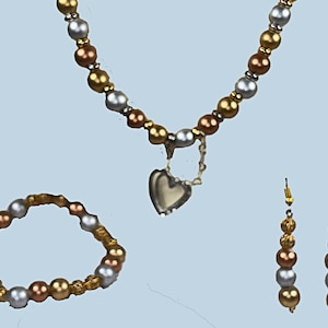 Può includere: Un set di gioielli composto da collana, bracciale e orecchini. La collana ha un ciondolo a forma di cuore ed è realizzata con perline dorate, argentate e color rame. Il bracciale e gli orecchini riprendono i colori delle perline. Sfondo azzurro chiaro.