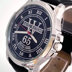 May include: A silver-toned wristwatch with a black face. The watch face features Roman numerals, the name "DAVID WATKINS", the number "65", and the date "05-02-1959". The watch has a black leather band.