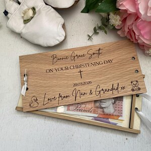 May include: A wooden christening gift box with engraved details, including a name, date, and message. The box is open, revealing banknotes. White baby shoes and pink flowers are in the background.
