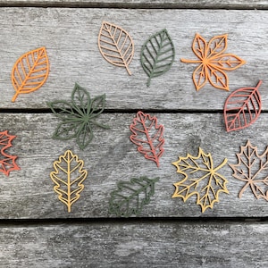May include: A collection of colourful, cutout leaf shapes in shades of orange, red, green, and yellow. The leaves are arranged on a weathered, grey wooden surface, suggesting an autumn theme. Various leaf types are visible.