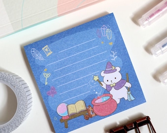 May include: Blue lined notepad with a cute cartoon bear wearing a witch hat and holding a wand. The bear is standing next to a cauldron and a book. There are stars, hearts, and crystals around the bear.