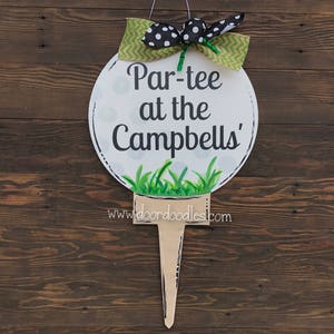 May include: A white golf ball door hanger with green grass and black polka dots. The text on the golf ball reads "Par-tee at the Campbells".