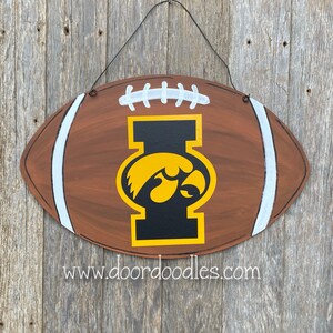 May include: A brown and white football-shaped door hanger with a black and gold University of Iowa logo. The logo features a black "I" with a yellow hawk inside. The text "www.doordoodles.com" is printed below the logo.