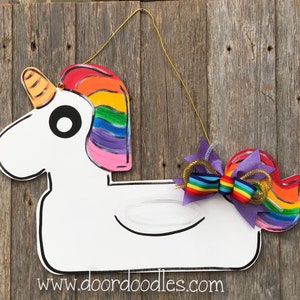 May include: A white unicorn door hanger with a rainbow mane and tail. The unicorn has a large black eye and a gold horn. The tail is decorated with a purple bow with a rainbow ribbon and gold glitter.