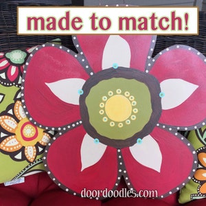 May include: A red, white, and green painted wooden flower with a brown center and white dots around the edge. The flower is sitting on a wicker chair with floral patterned cushions. The text "made to match!" is above the flower and the text "doordoodles.com" is below the flower.