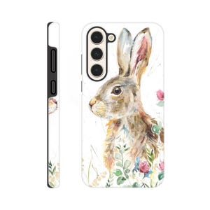 May include: A white phone case featuring a watercolor painting of a brown and tan rabbit with long ears, surrounded by green foliage and pink flowers. The side view shows a black border and camera cutouts.