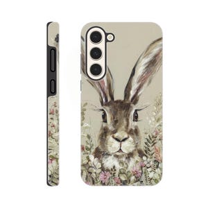 May include: A phone case featuring a detailed illustration of a brown rabbit with long ears, surrounded by wildflowers. The case has a neutral background and a black side profile. The design is artistic and nature-themed.