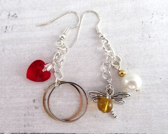 May include: Silver dangle earrings. One earring features a red heart-shaped crystal and two silver hoops. The other earring has a dragonfly charm with a yellow bead, a white pearl, and a gold bead. The earrings have silver chain accents.