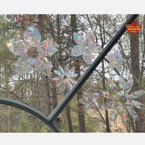 May include: A set of iridescent flower window clings, made from recycled materials, are displayed on a window. The clings are shaped like flowers with multiple petals and a center circle. The background is a blurry image of trees and a blue sky.