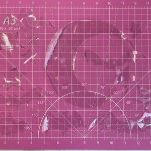 May include: A pink cutting mat with a grid pattern and a semi-circle protractor. The mat is labeled "A3 (45 x 30 cm)". There are pieces of clear plastic on the mat.
