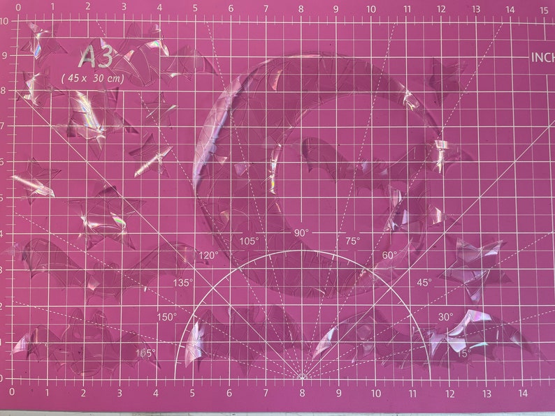 May include: A pink cutting mat with a grid pattern and a semi-circle protractor. The mat is labeled "A3 (45 x 30 cm)". There are pieces of clear plastic on the mat.