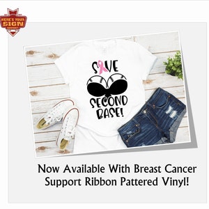May include: White t-shirt with a pink ribbon and black baseballs with the text "Save Second Base!" printed on it. The shirt is laid out on a wooden surface with a pair of white sneakers and a pair of blue denim shorts. The text "Now Available With Breast Cancer Support Ribbon Pattered Vinyl!" is printed below the image.