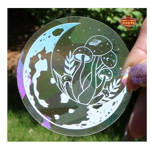 May include: A clear iridescent sticker with a white crescent moon and mushrooms with a planet and stars inside the moon.