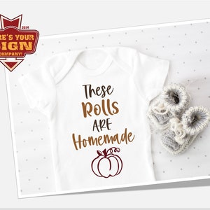 May include: White baby onesie with the text "These rolls are homemade" and a brown outline of a pumpkin with a red interior.