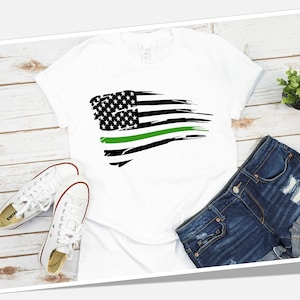 May include: A white t-shirt with a distressed American flag design featuring a green stripe. The flag is on a white background. The t-shirt is laying on a wooden surface with a pair of white sneakers and a pair of blue denim shorts.