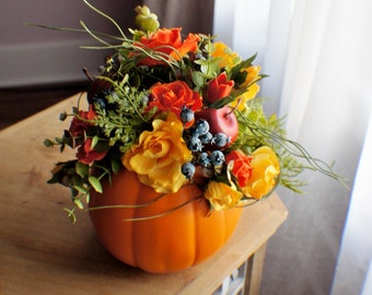 May include: An orange pumpkin-shaped container overflowing with a vibrant floral arrangement. The arrangement includes orange and yellow roses, red berries, and green foliage, creating a festive autumn centerpiece. The pumpkin sits on a wooden table.