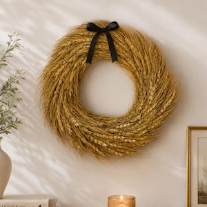 May include: A wheat wreath with a black bow hangs on a white wall. A vase with greenery, books, a lit candle in a glass holder, and a wooden bowl with beads sit on a wooden table. The books are titled "THE KINFOLK", "LIVE BEAUTIFUL", and "THIS IS HOME".