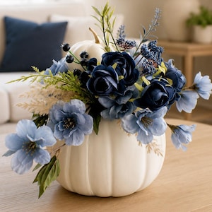 Peut inclure: Une citrouille décorative blanche servant de centre de table, agrémentée d'un arrangement de fleurs artificielles bleu foncé et bleu clair, de petites baies sombres et d'un feuillage vert fin. Les fleurs sont disposées sur le dessus de la citrouille.