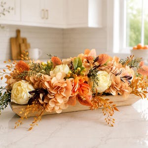 May include: A long, rectangular wooden dough bowl filled with an arrangement of artificial flowers in shades of cream, peach, and burnt orange, accented with green foliage, small berries, and dried-style stems extending from the sides.