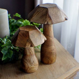 May include: Two wooden mushroom sculptures, one larger than the other, with a brown, natural wood finish. The mushrooms are sitting on a wooden table.