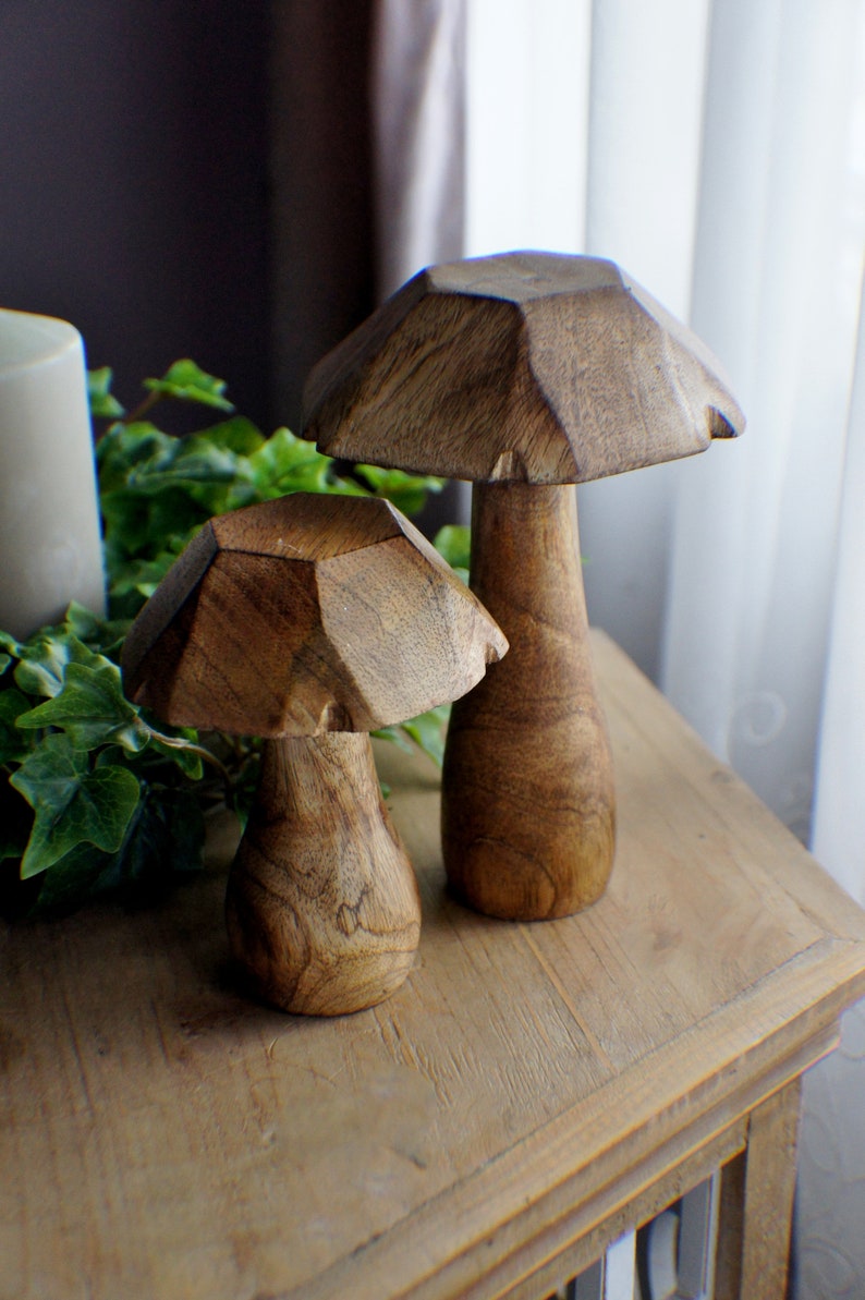 May include: Two wooden mushroom sculptures, one larger than the other, with a brown, natural wood finish. The mushrooms are sitting on a wooden table.